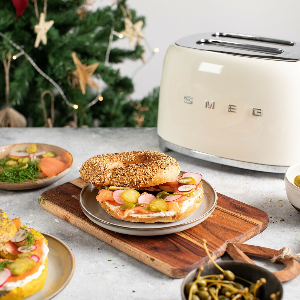 Three bagel halves topped with cream cheese, smoked salmon, and dill, arranged on a board with festive greenery.