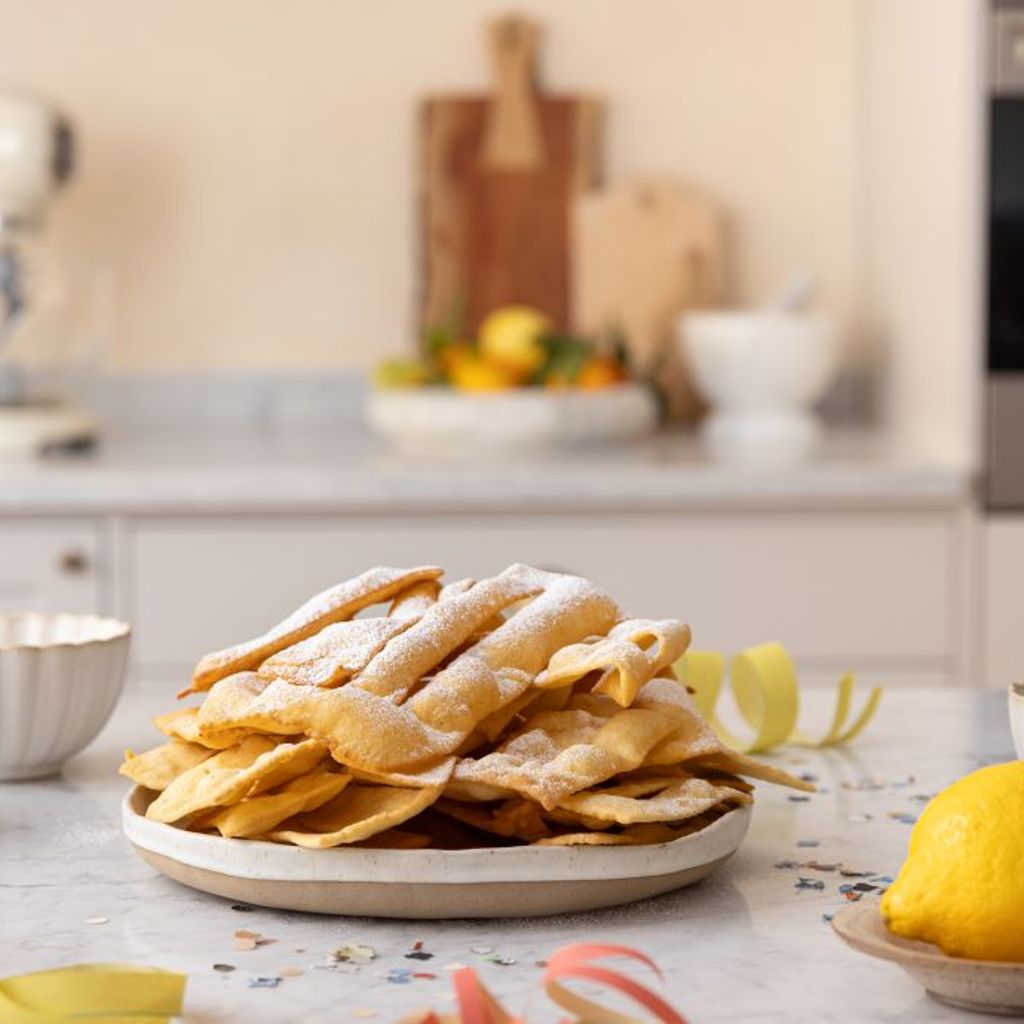 A tray of crispy chiacchiere dusted with icing sugar on a marble worktop, with a lemon and ingredients in the Smeg kitchen.