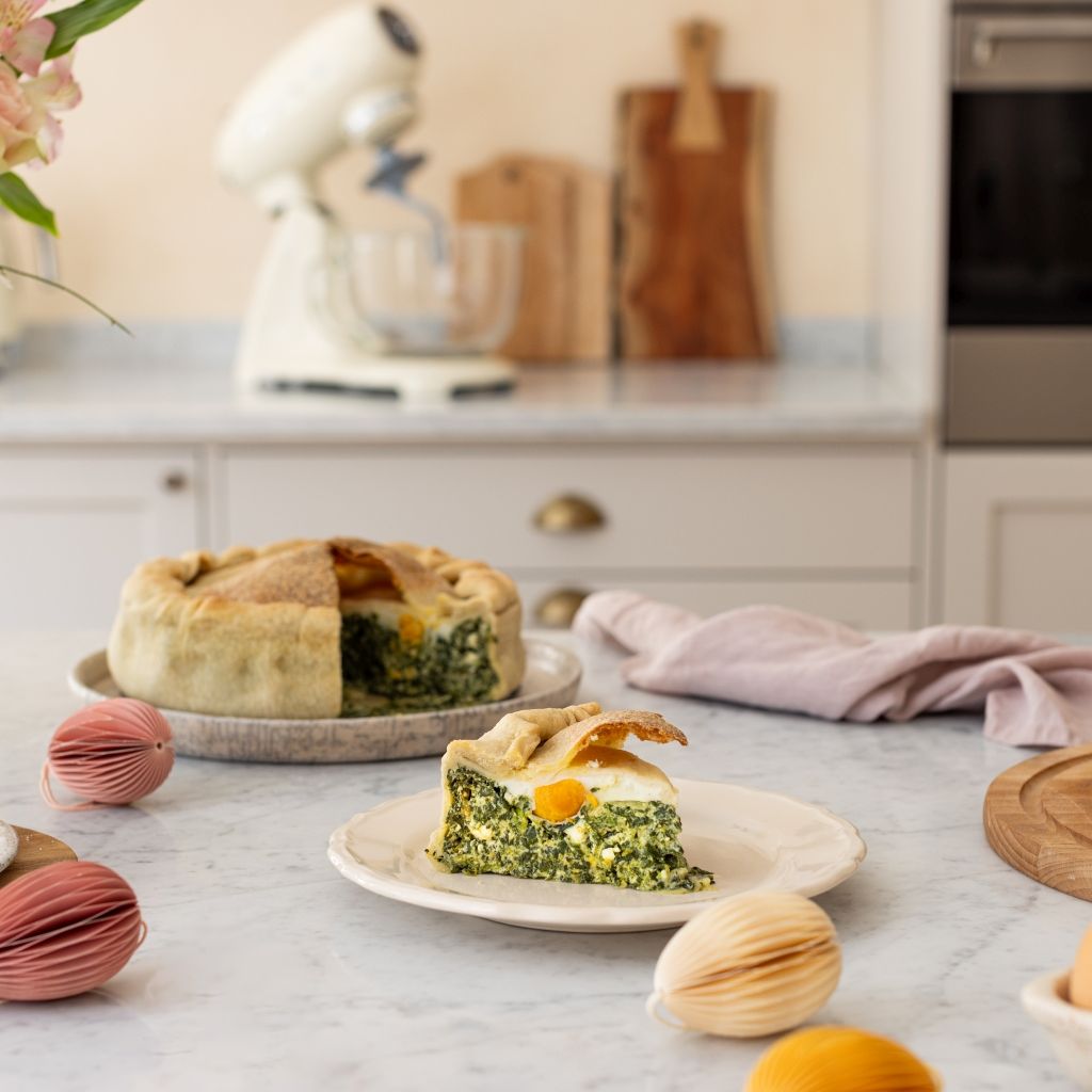 A slice of torta pasqualina filled with spinach, eggs, and ricotta on a plate, with the whole cake, Easter decorations, and a Smeg stand mixer in the background.