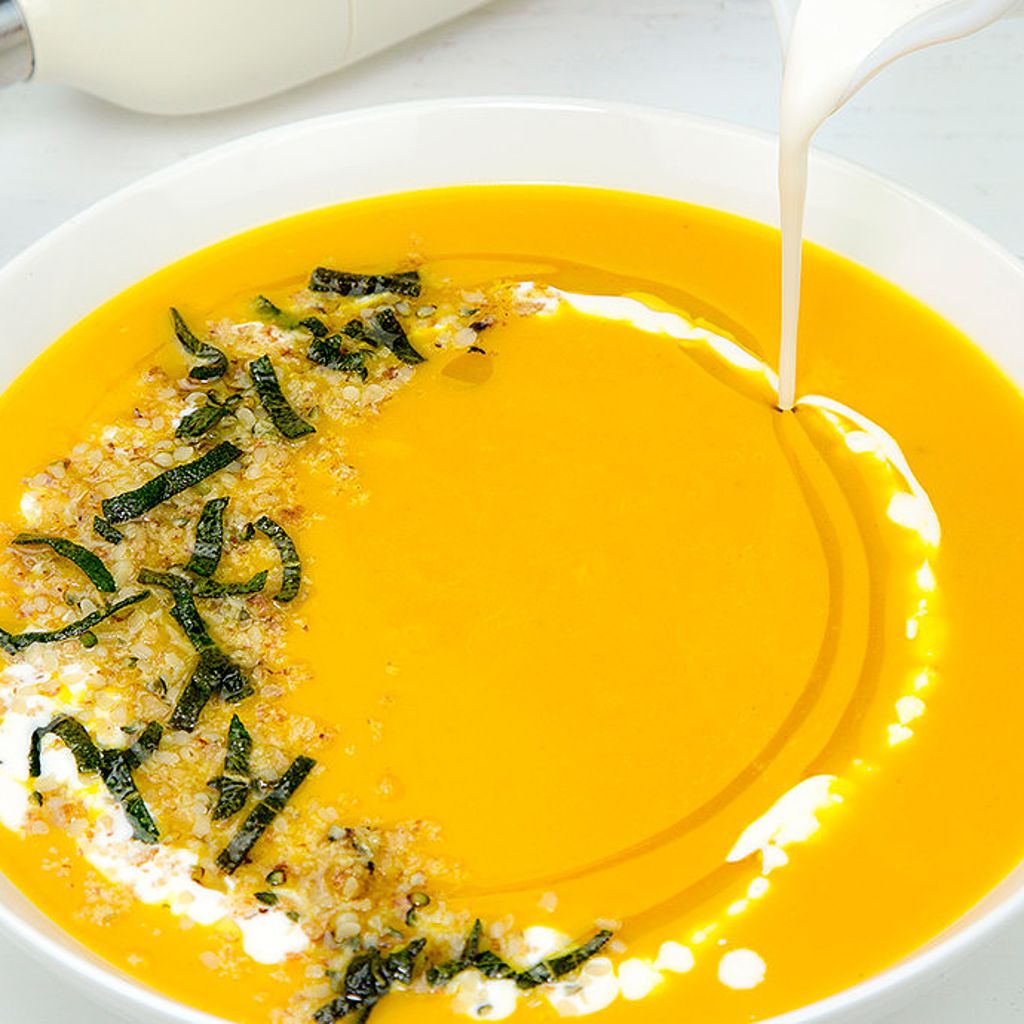 Cream being poured into a bowl of bright orange pumpkin soup topped with herbs and grains.