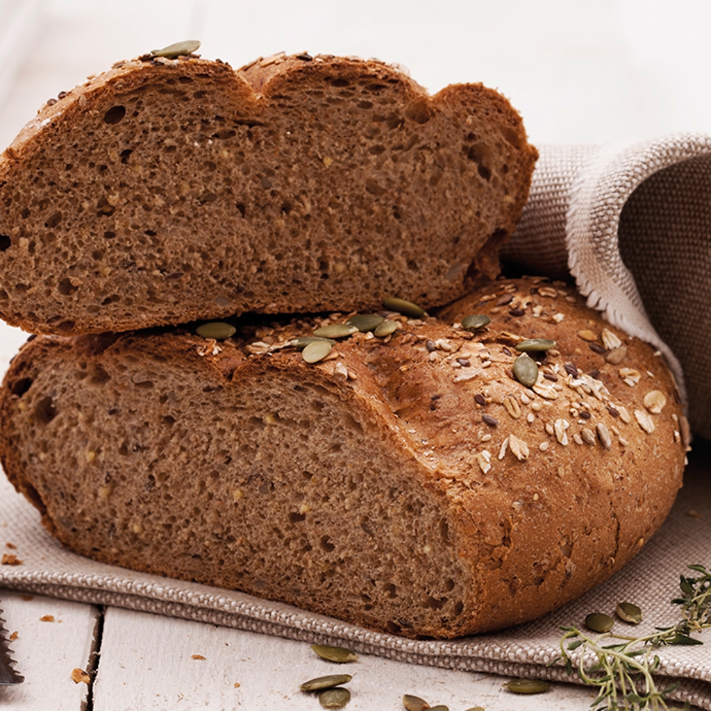 A sliced loaf of dark rye bread topped with pumpkin and sunflower seeds, resting on a linen cloth next to a bread knife.