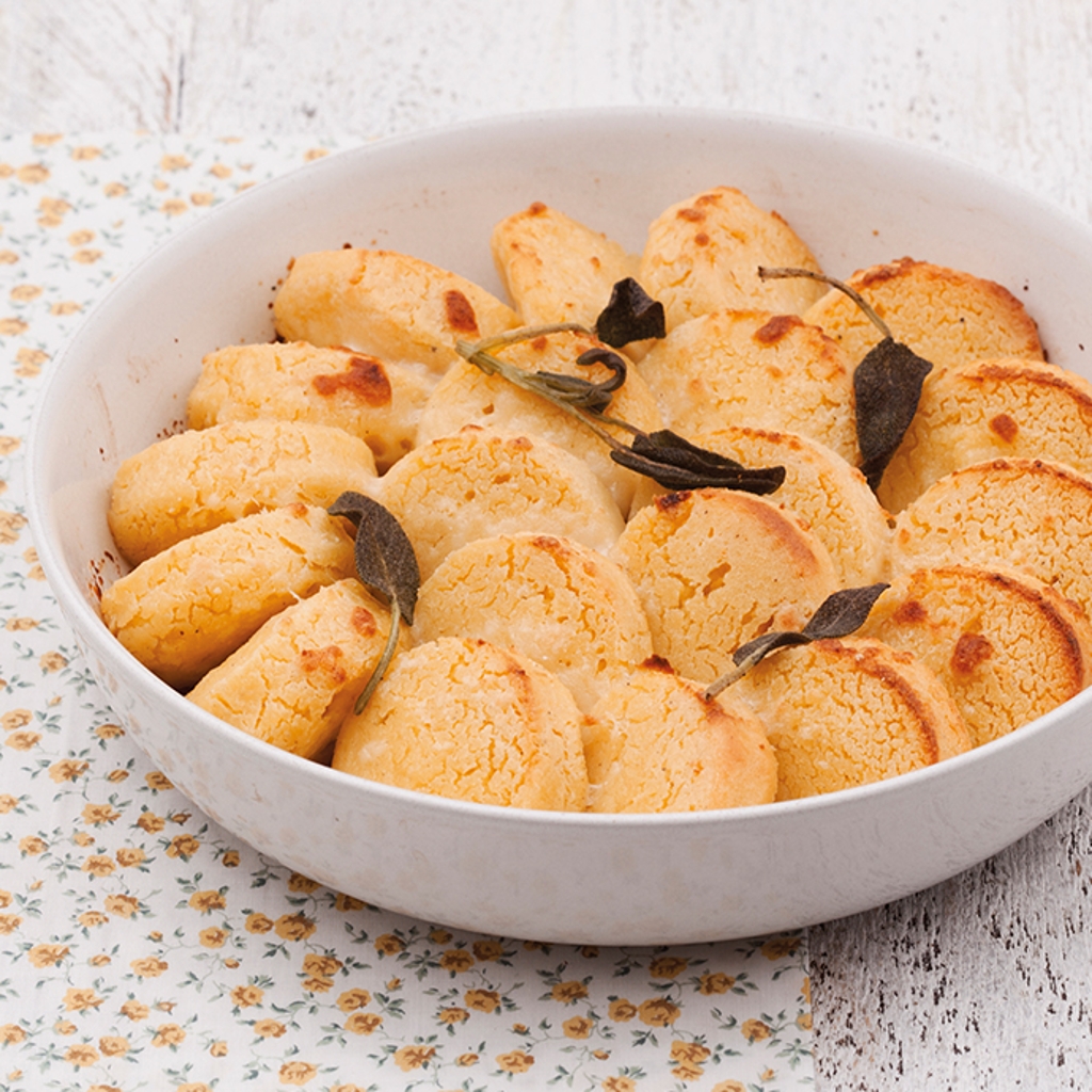 Golden-brown, circular semolina gnocchi baked in a white dish, garnished with crisp sage leaves on a floral tablecloth.