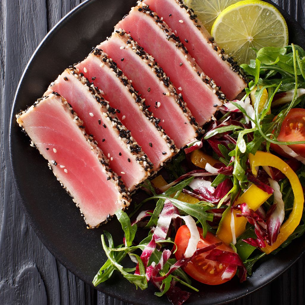 Slices of rare, pink seared tuna with a sesame crust served with a fresh garden salad and lemon on a black plate.