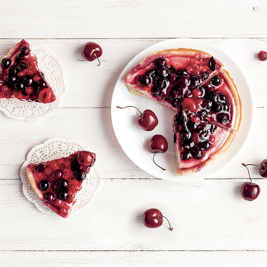 Overhead view of a berry-topped cheesecake on a white wooden table, with loose cherries and individual slices on lace doilies.