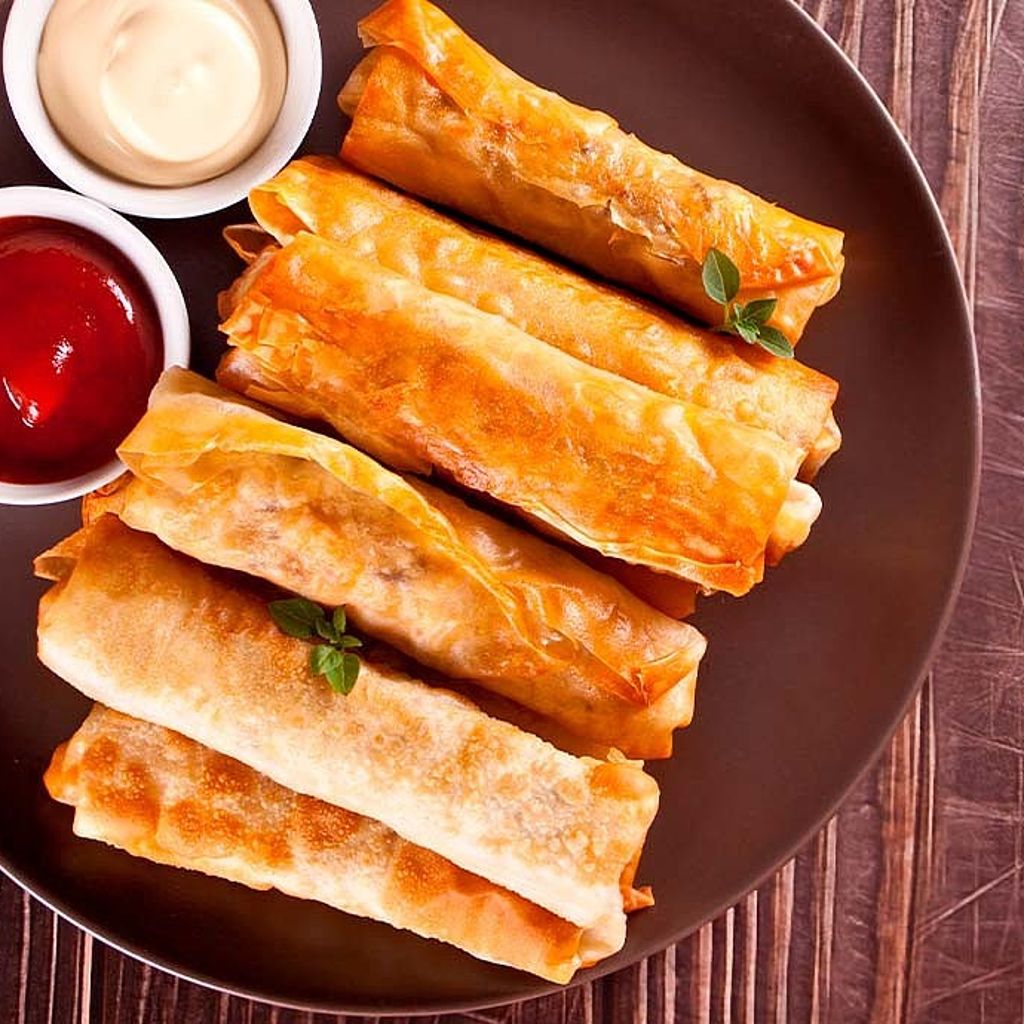 Five golden-brown, crispy spring rolls on a dark plate served with side bowls of red and white dipping sauces.