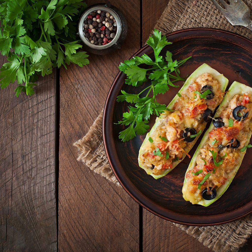 Top-down view of two zucchini halves stuffed with a tofu and vegetable mixture, topped with melted cheese and olives.