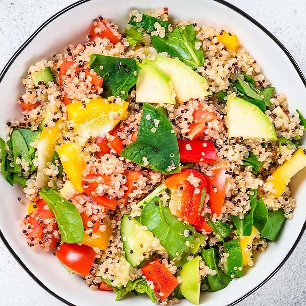 Overhead view of a vibrant quinoa salad mixed with chopped red and yellow peppers, cucumber, and spinach in a white bowl.