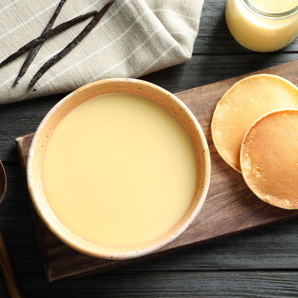 Overhead view of a bowl of smooth yellow custard cream on a wooden board with small pancakes and vanilla beans.