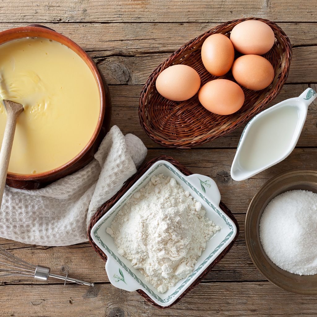Overhead view of eggs in a basket, a bowl of flour, milk, and cream base on a rustic wooden table.