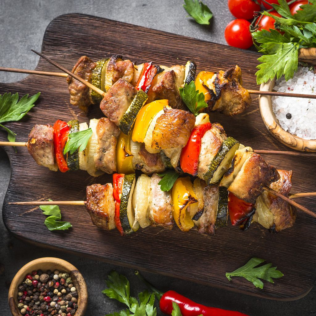 Three grilled skewers with pieces of meat, zucchini, onions, and bell peppers on a dark wooden board.