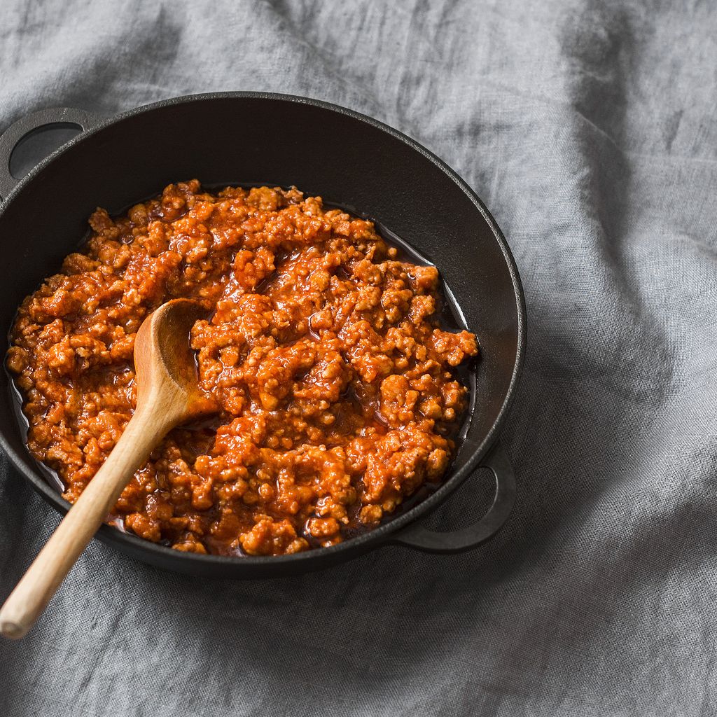 Thick, savory beef ragu sauce being stirred with a wooden spoon in a black cast iron pan on a grey linen cloth.