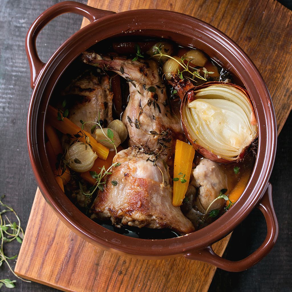 Steamed rabbit pieces in a brown ceramic pot with carrots, onions, and fresh thyme, served on a wooden board.