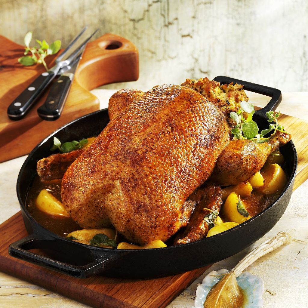 Whole golden-brown roasted duck in a black cast-iron pan, served with savory stuffing and sliced apples in a light sauce.