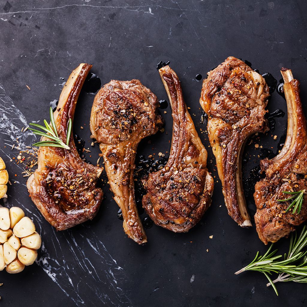 Four grilled lamb loin chops seasoned with cracked pepper and spices, garnished with rosemary on a dark marble surface.