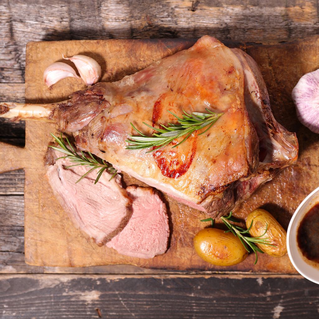 Sliced roasted leg of lamb on a rustic wooden board, garnished with fresh rosemary sprigs, garlic, and small potatoes.
