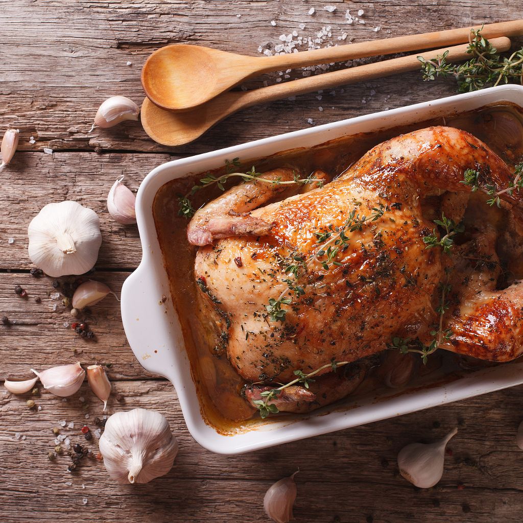 Whole golden-brown roasted chicken in a white baking dish, seasoned with herbs and surrounded by fresh garlic cloves.
