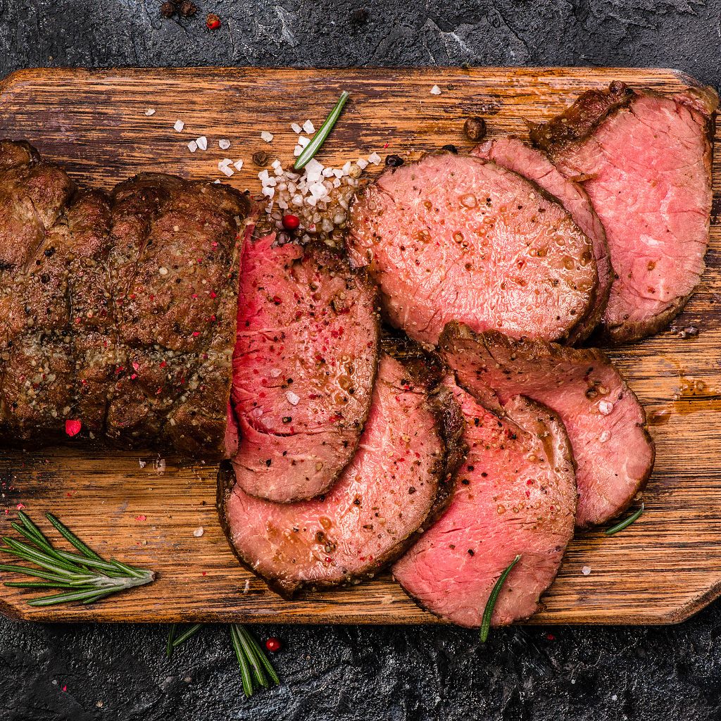 Sliced medium-rare roast beef on a wooden board, seasoned with sea salt, peppercorns, and fresh rosemary.