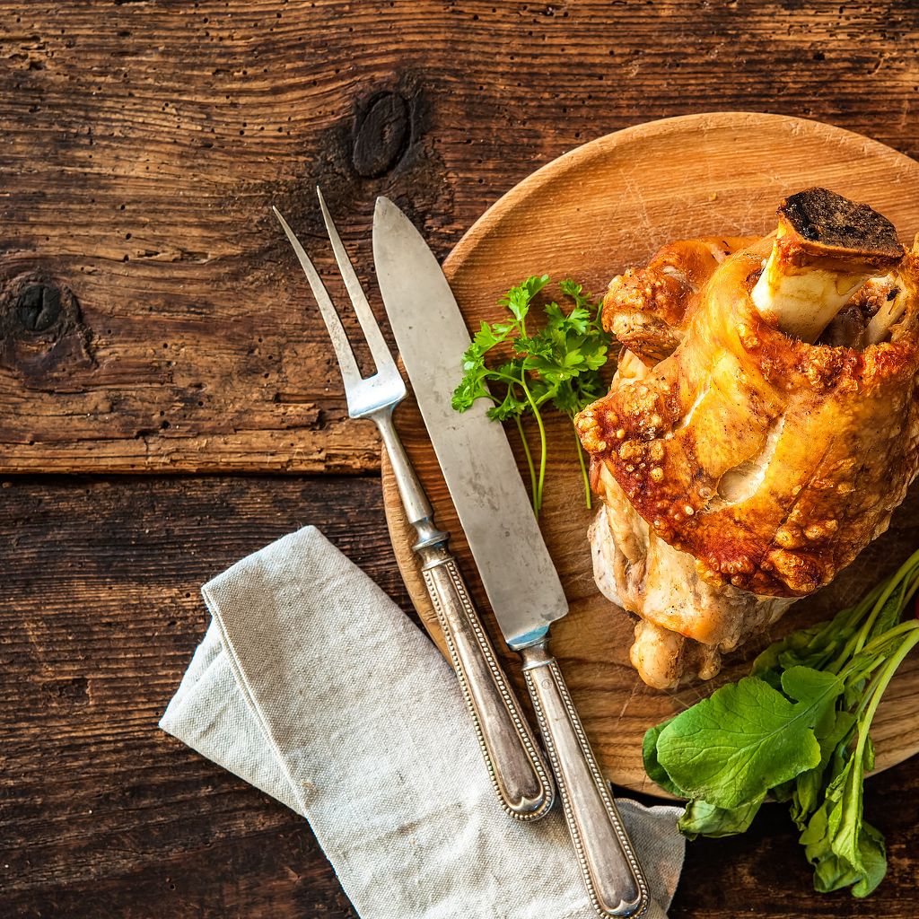 Crispy roasted pork shank on a wooden board with a vintage carving fork and knife, garnished with fresh parsley.
