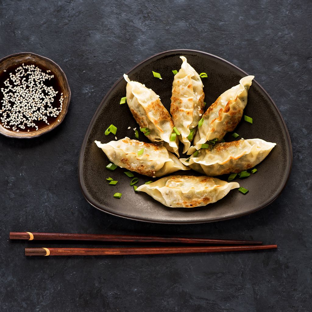 Six golden-brown pan-fried dumplings on a dark plate, garnished with green onions, sesame seeds, and dipping sauce.