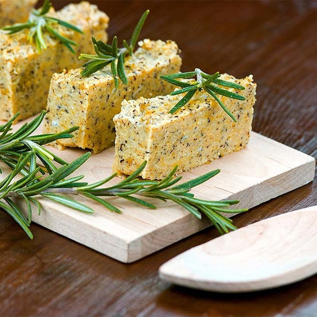 Rectangular slices of golden herbed polenta topped with rosemary sprigs on a light wood board.