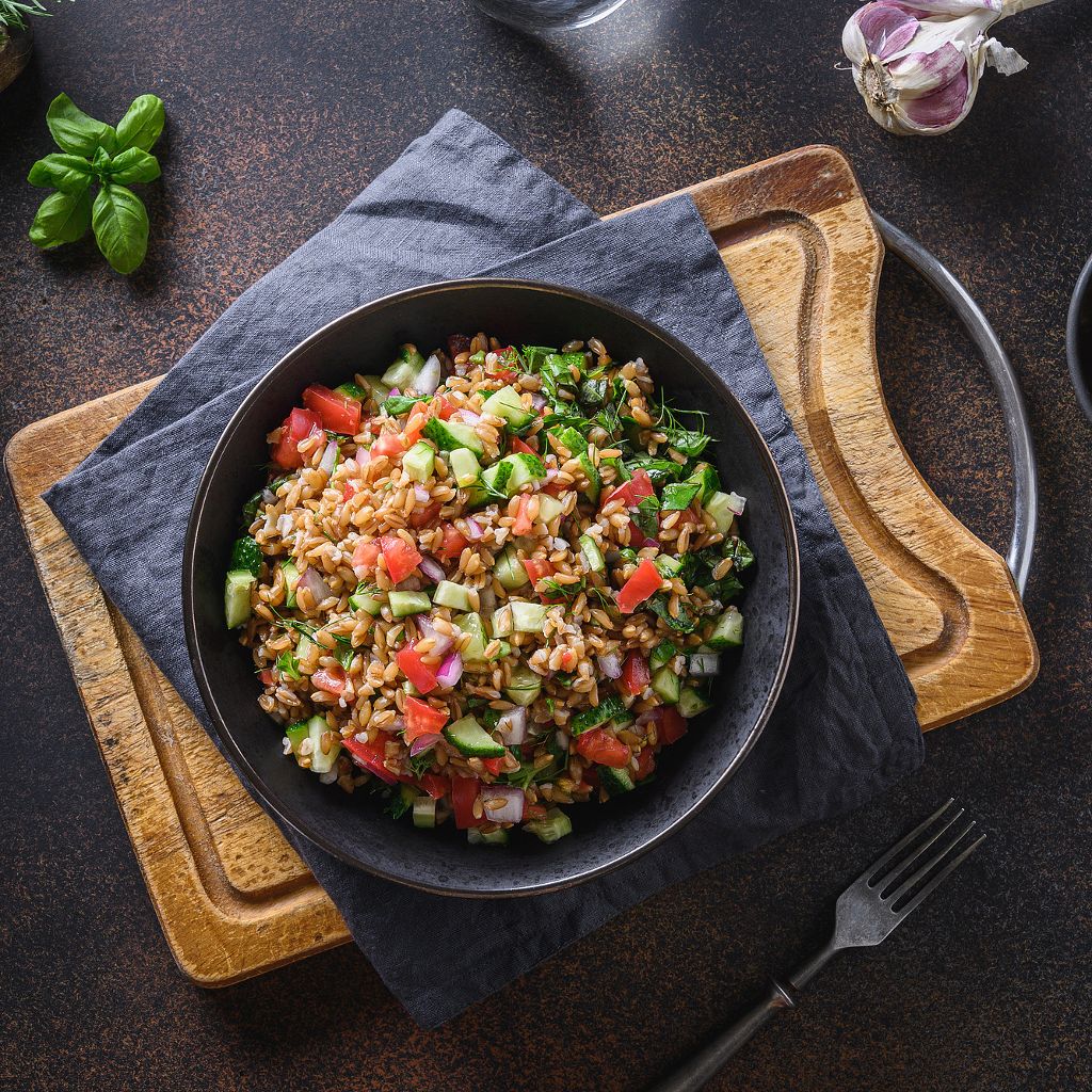 Top-down view of a dark bowl filled with spelt, diced tomatoes, cucumbers, and red onions on a rustic wooden surface.