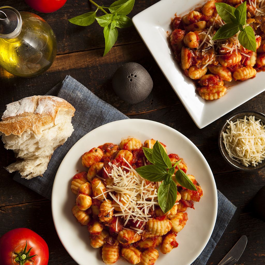 Plated potato gnocchi topped with tomato sauce, shredded cheese, and basil on a dark wood table with fresh tomatoes.