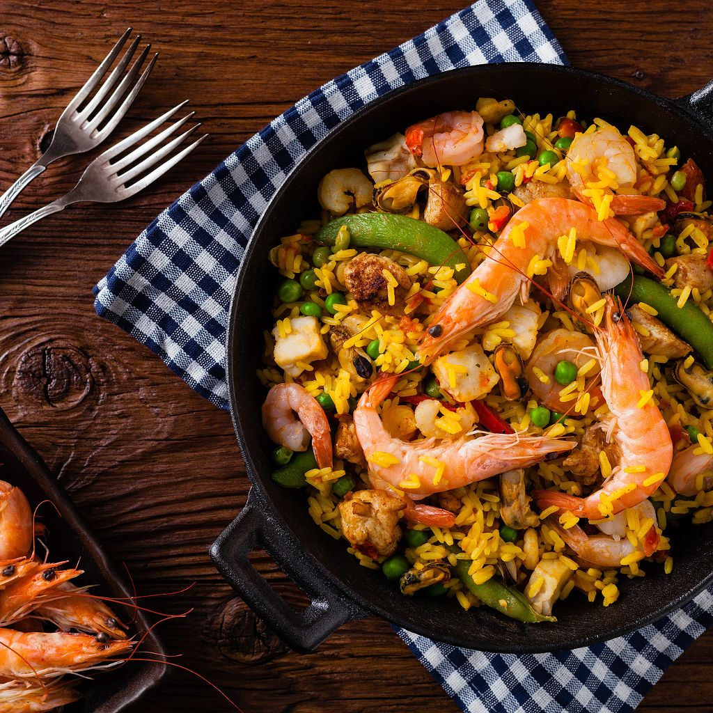 Baked paella pan with saffron rice, shrimp, mussels, vegetables, and legumes, served on a rustic table with a checkered placemat.