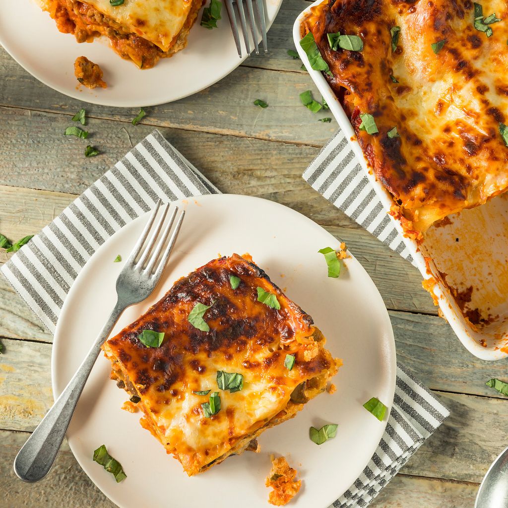 Squares of lasagna with golden melted cheese and fresh basil on white plates next to a full baking dish.