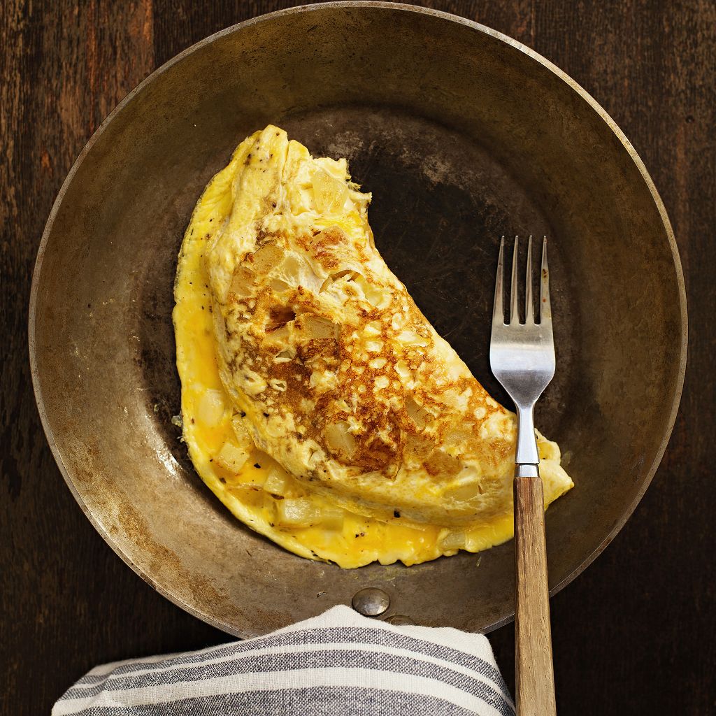 A folded golden-brown omelette with potato chunks in a rustic metal skillet, served with a wooden-handled fork.