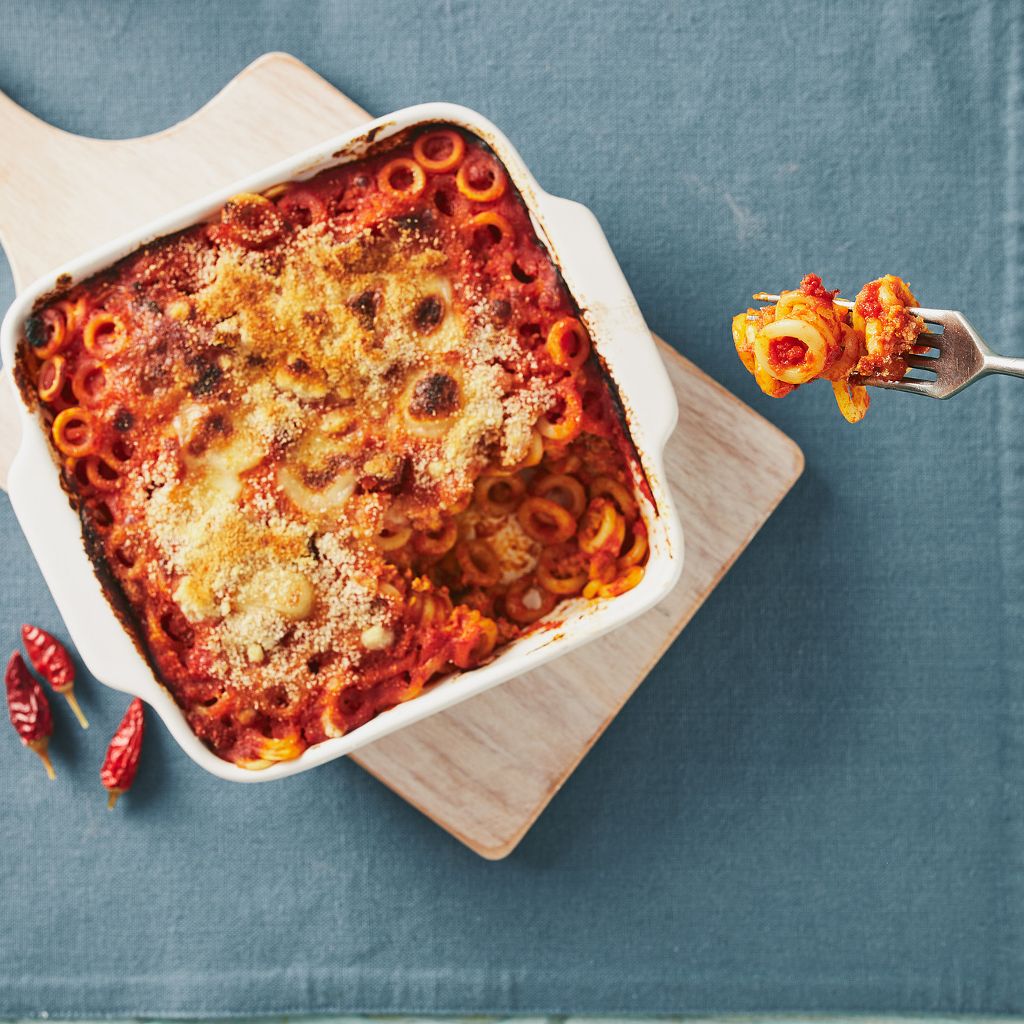 Ring-shaped pasta in red sauce with a toasted breadcrumb crust in a white dish, with a forkful being lifted.