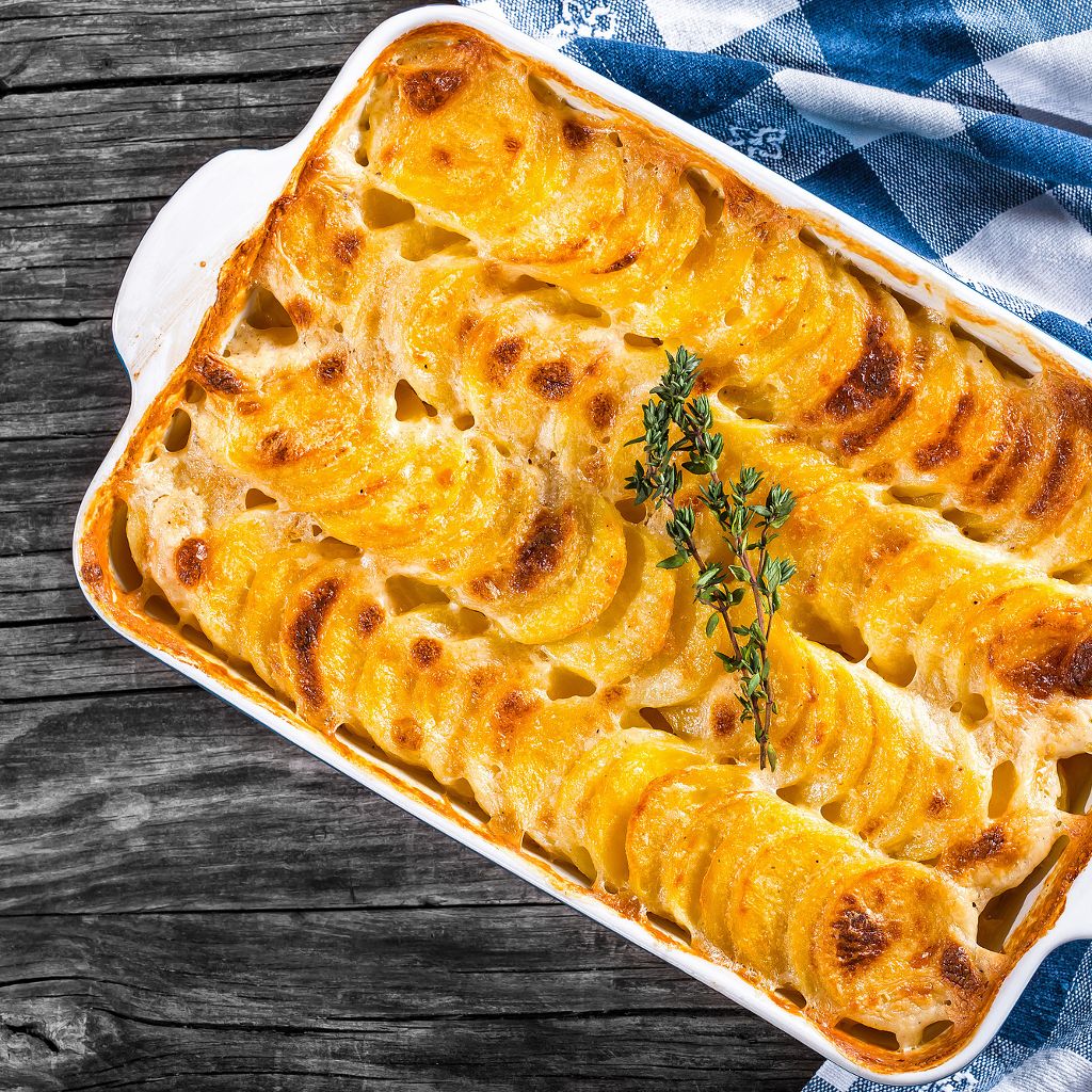 Sliced potatoes layered in a white dish, baked golden-brown with a rosemary sprig, on a dark wood background.