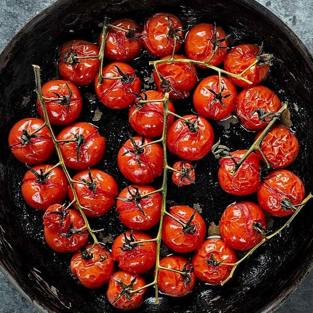 Top-down view of roasted red cherry tomatoes on the vine in a black skillet against a grey stone surface.