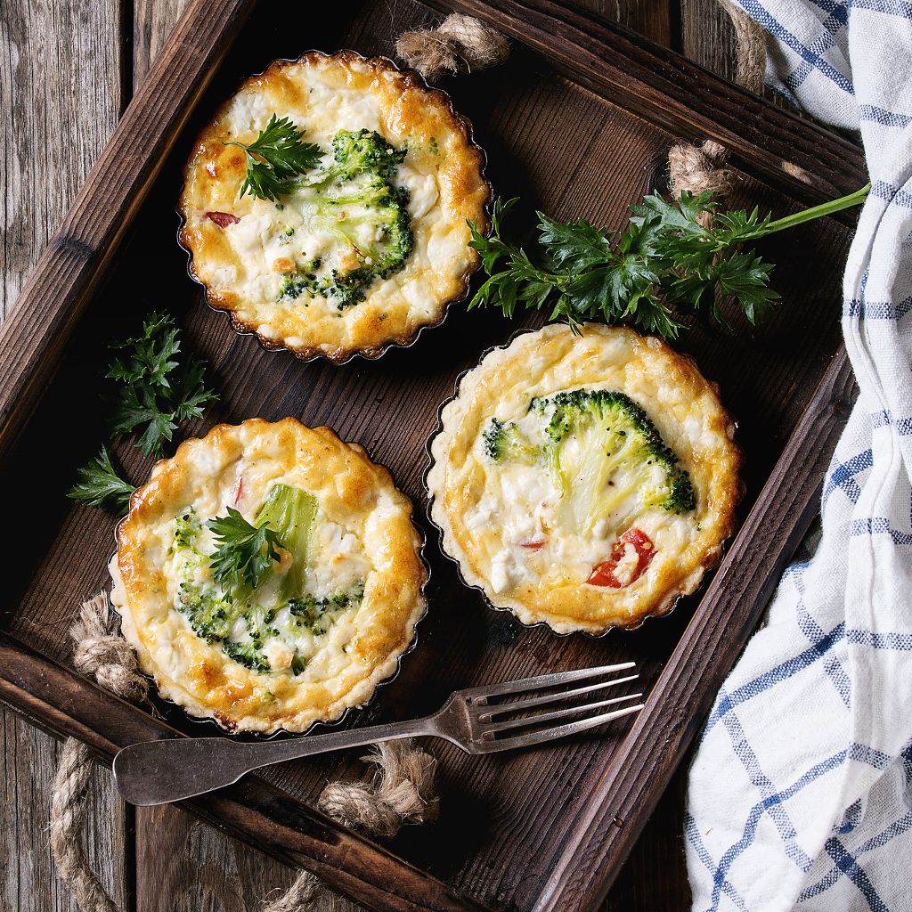 Three golden mini flans with broccoli florets in a wooden tray, garnished with fresh parsley and a silver fork nearby.