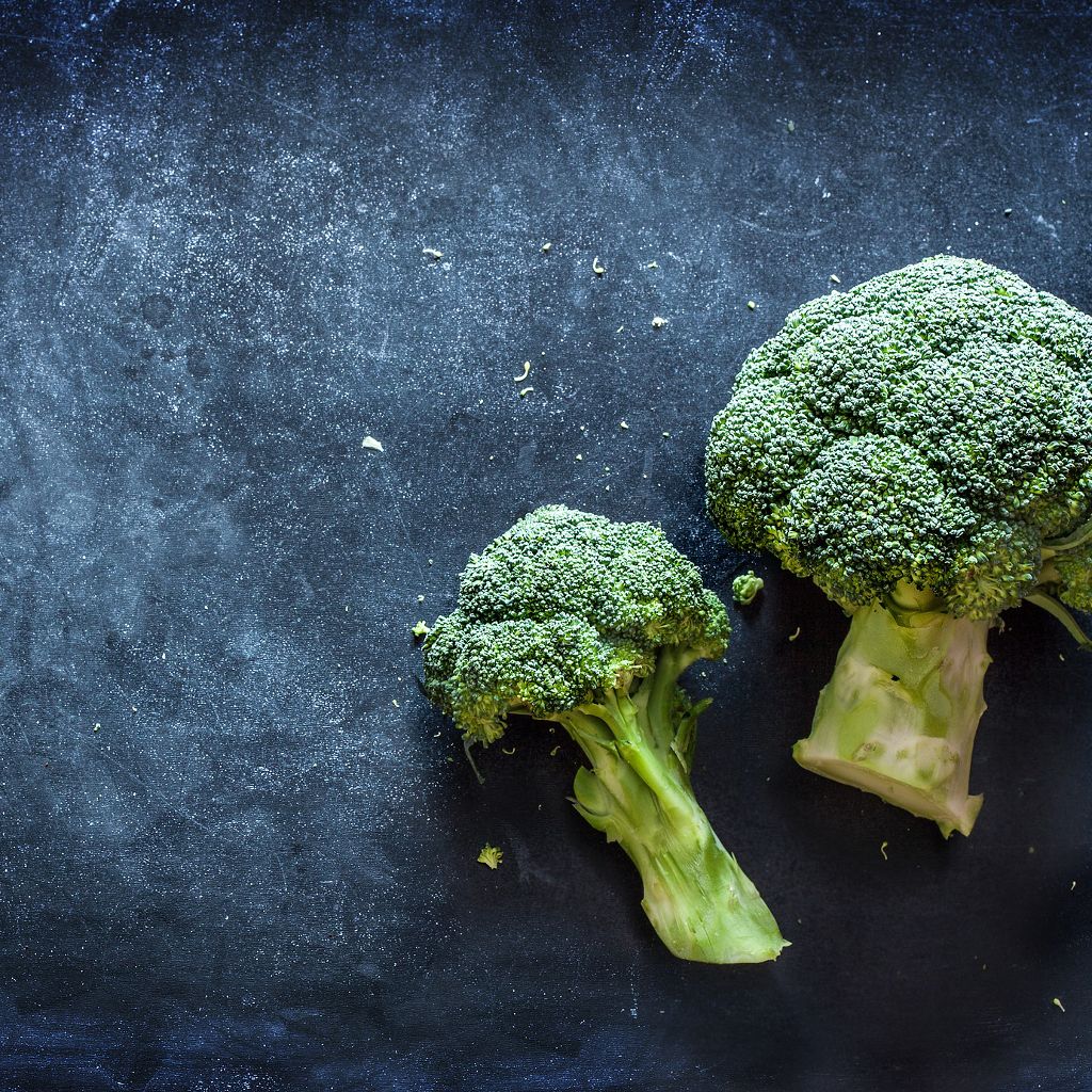 Two fresh green broccoli florets on a dark, textured blue surface with small crumbs.