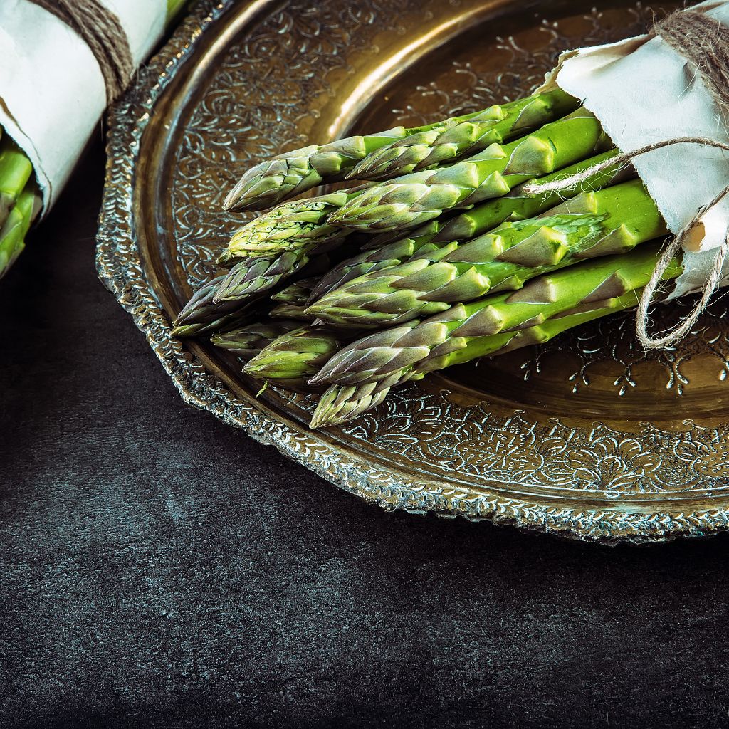 Vibrant green asparagus spears bundled in paper on an ornate brass platter over a dark textured surface.
