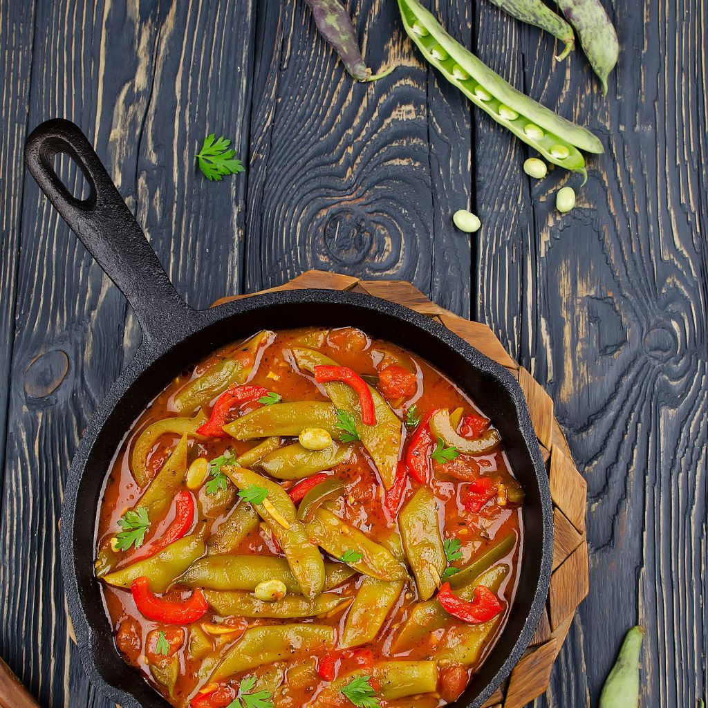 A black skillet of red pepper and green bean stew in tomato sauce, with raw beans on a dark wood table.