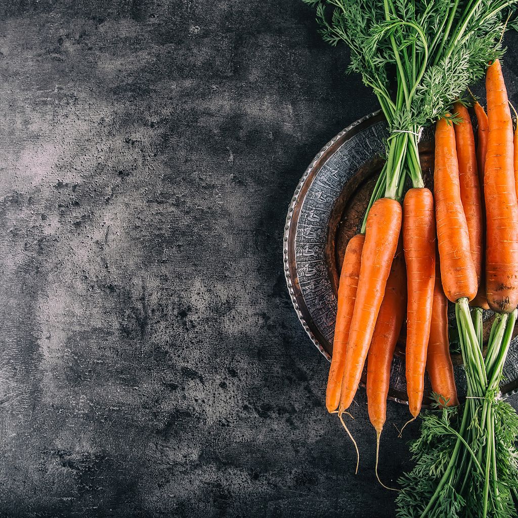 Bright orange carrots with leafy green stems resting on a dark metal plate against a textured charcoal background.