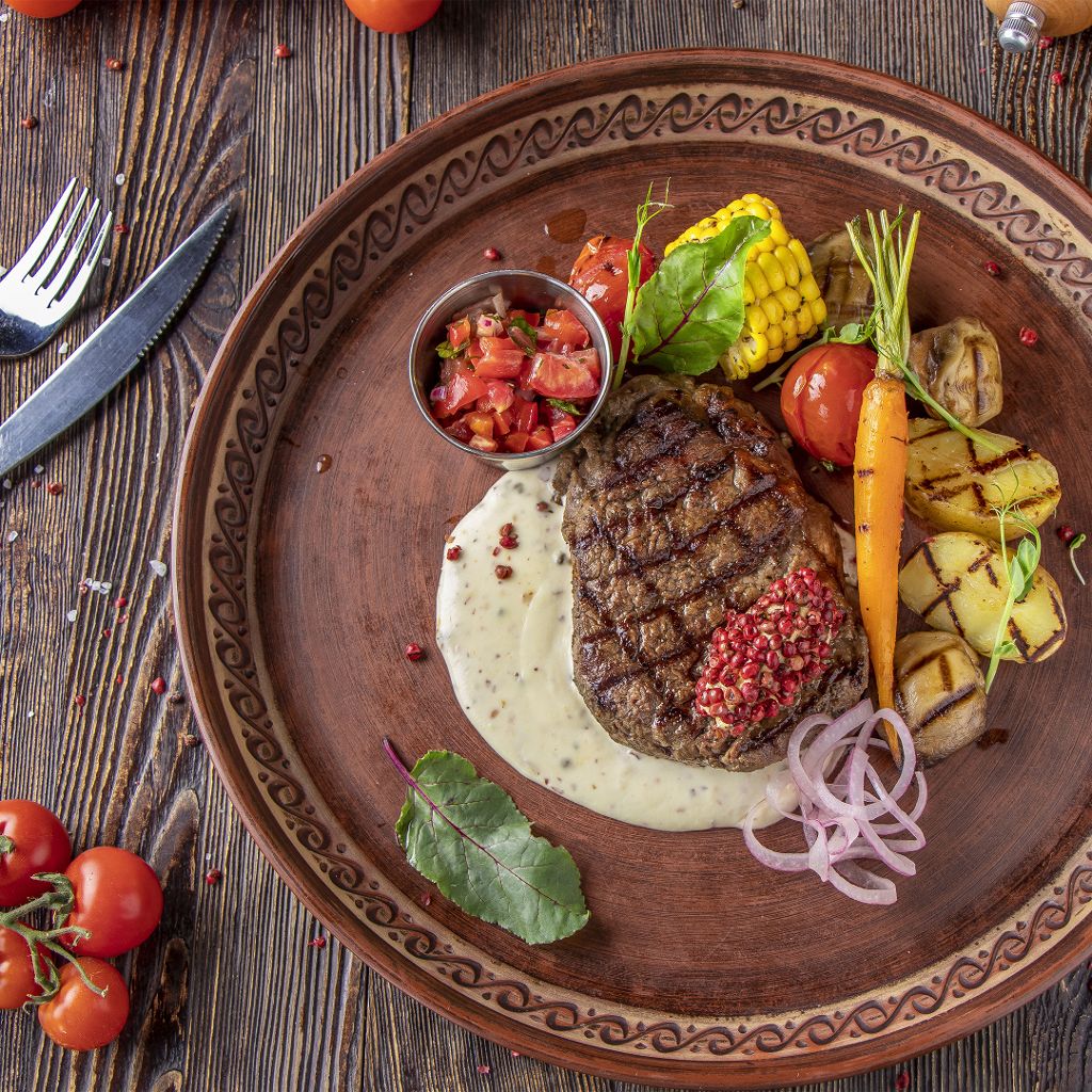 A grilled steak with crosshatch char marks, served with creamy sauce, pink peppercorns, corn, carrot, and tomato.