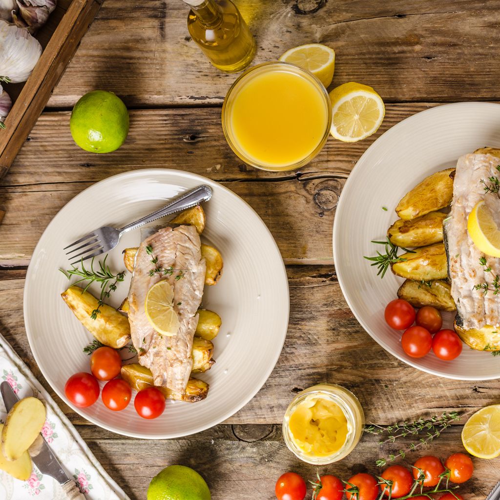 A top-down view of a roasted fish fillet over potato wedges and cherry tomatoes, garnished with lemon and rosemary.