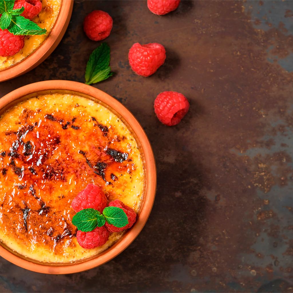 Two terracotta bowls of custard with a caramelized sugar topping, garnished with fresh raspberries and mint leaves.