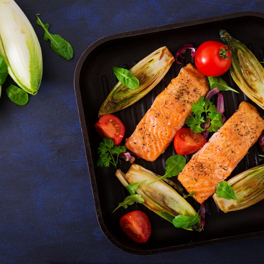 Grilled salmon fillets in a ribbed pan with charred chicory, halved cherry tomatoes, and fresh herbs.