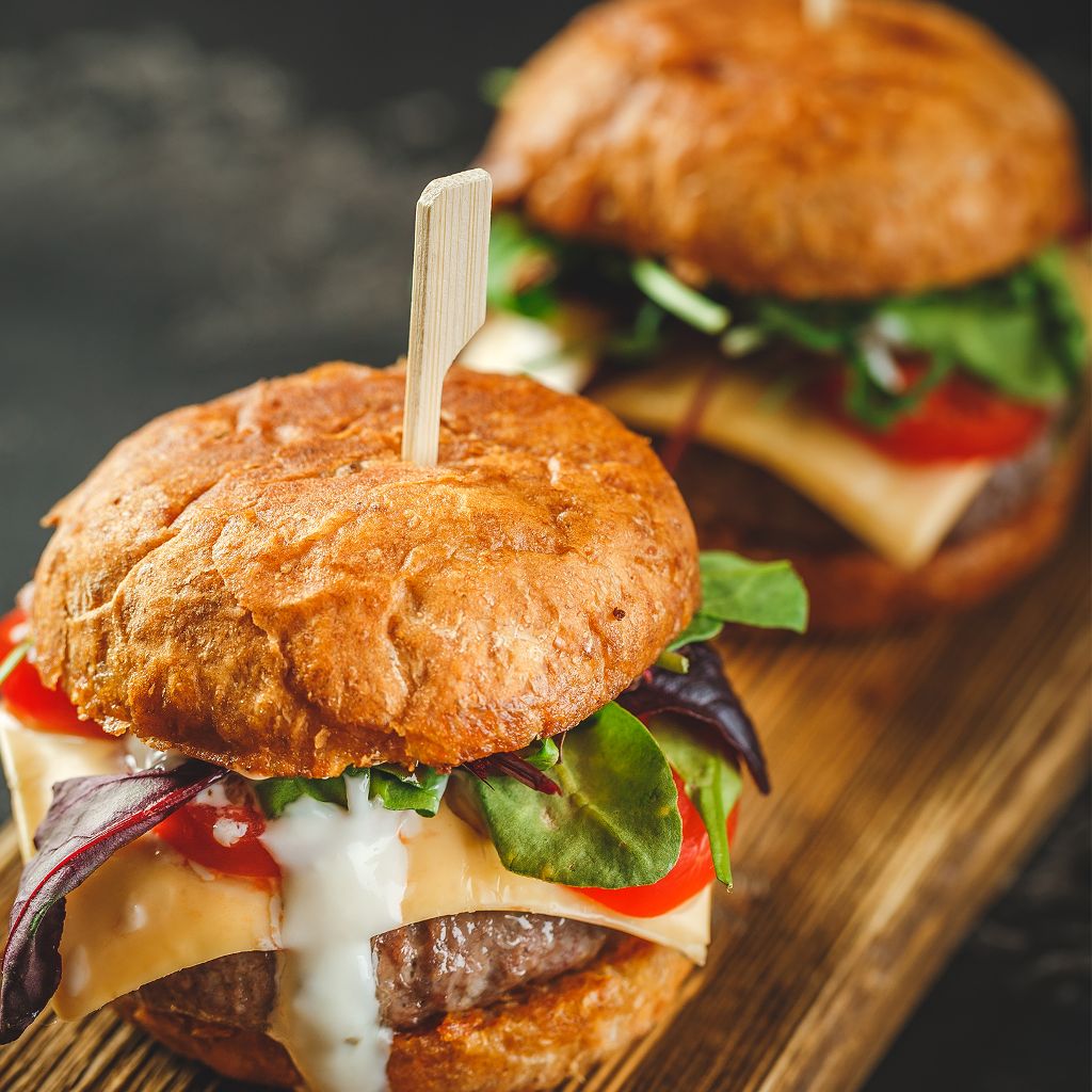 Two thick beef burgers on toasted buns with melted cheese, lettuce, tomato, and a creamy white sauce.