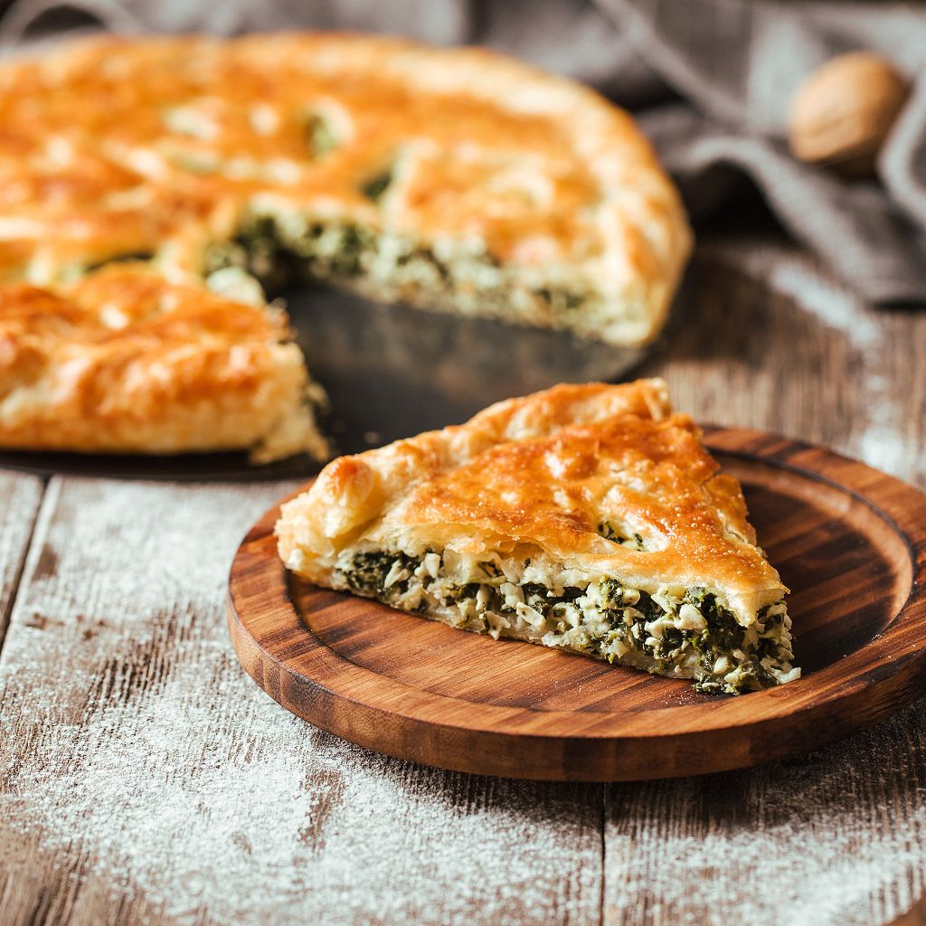 A slice of golden-brown flaky pastry filled with greens and cheese on a wood plate, with the whole pie behind it.