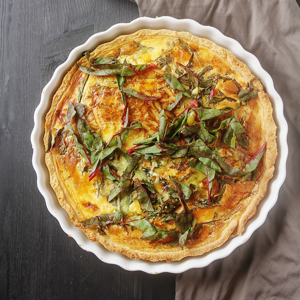 Top view of a golden savory pie in a white fluted dish, topped with fresh leafy greens on a dark wooden surface.