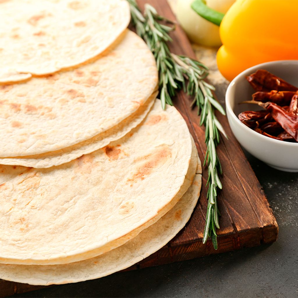Three soft, round piadinas stacked on a wooden board with small bowls of toppings and fresh herbs.