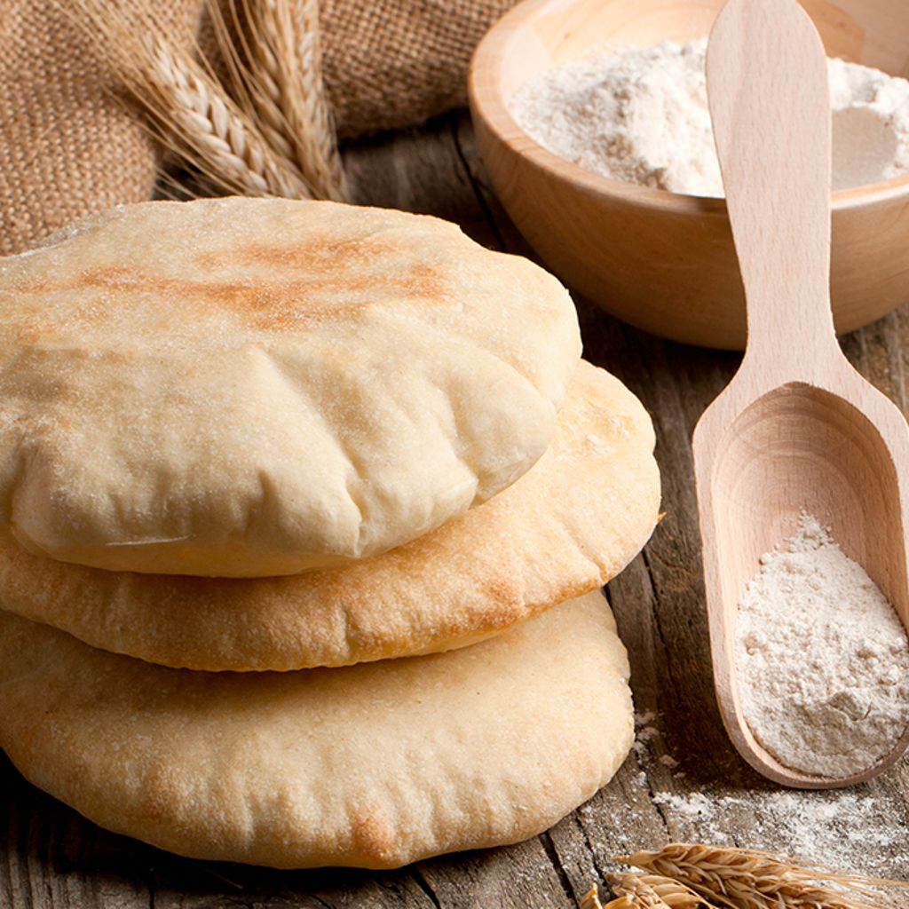 A stack of three soft pita breads on a wooden surface with a wooden scoop of flour and a bowl in the background.