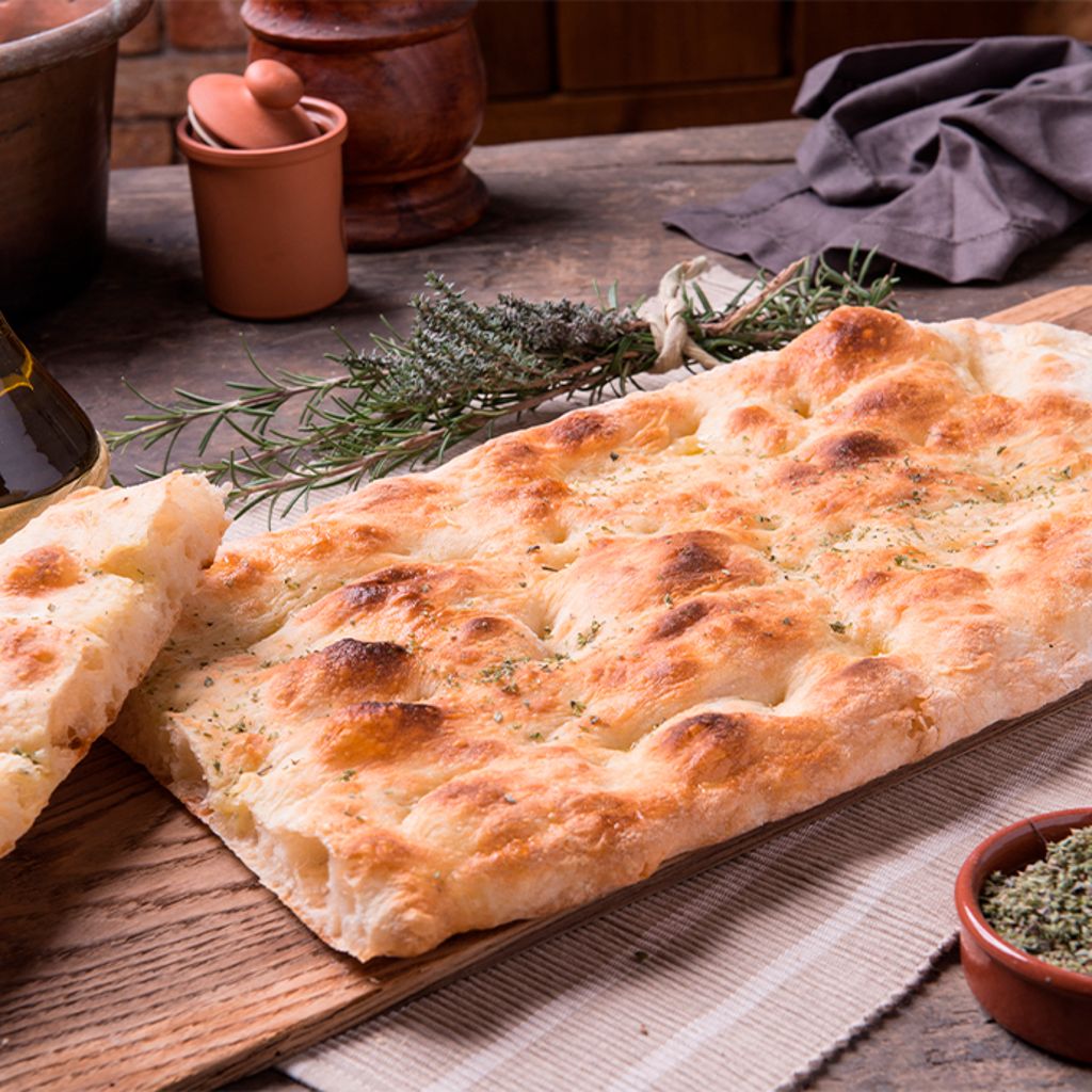 Golden-brown flatbread on a wooden board with olive oil, rosemary sprigs, and a bowl of dried herbs.