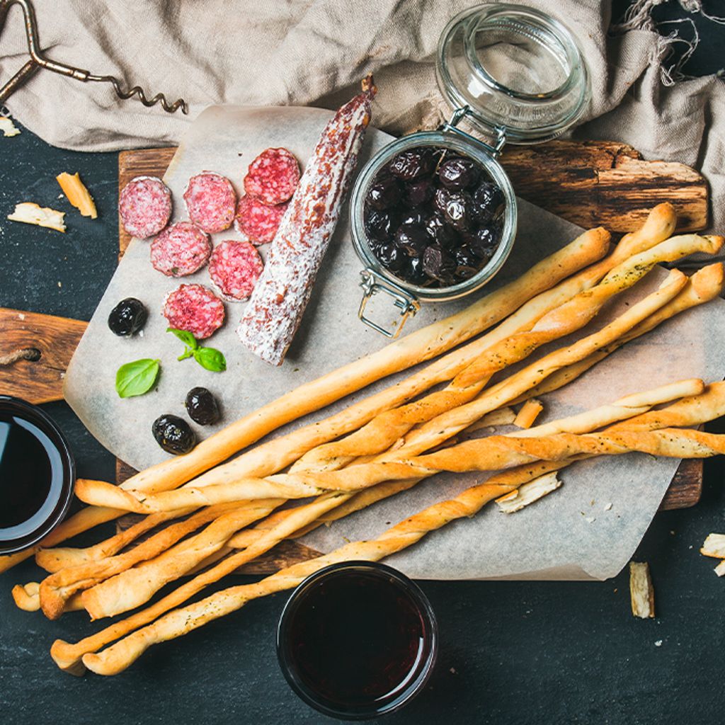 Long breadsticks, sliced salami, and a jar of black olives on a wooden board with two glasses of red wine