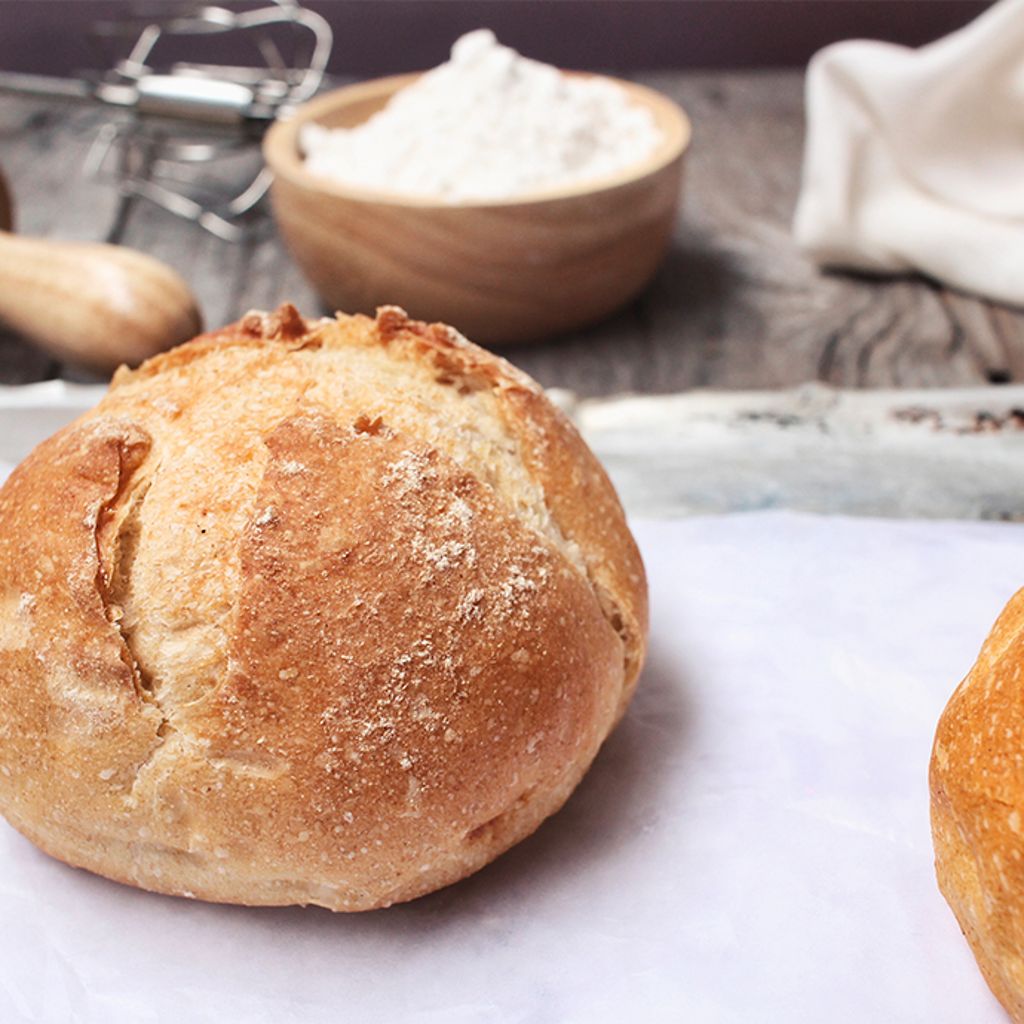 Two golden-brown crusty bread loaves on parchment paper with a rolling pin and flour bowl in the background.
