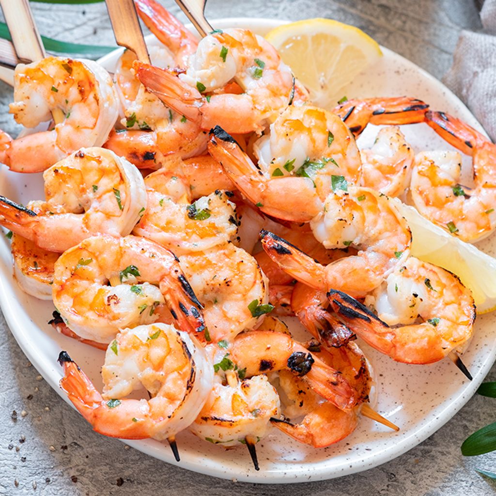 Three grilled prawn skewers garnished with fresh herbs and lemon wedges on a dark, textured plate.